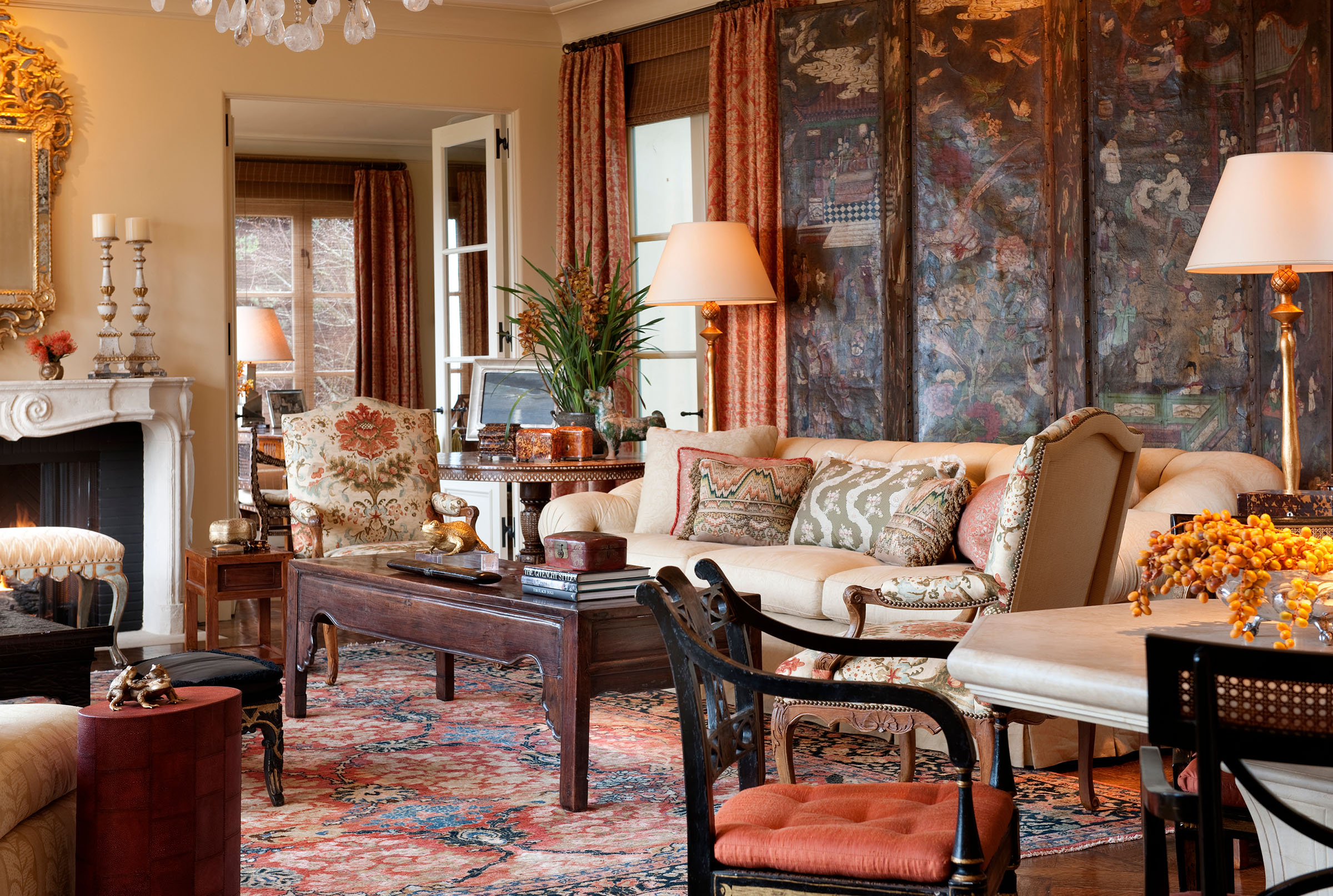 Elegant living room with ornate sofa, patterned cushions, antique wooden coffee table, Asian-style divider, and armchairs. Warm lighting, detailed rug, fireplace with candles, mirrors, and decorative items create a cozy, classic atmosphere reminiscent of a richly decorated Mediterranean home.